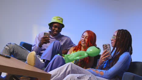 Studio-Shot-Of-Group-Of-Young-Gen-Z-Friends-Sitting-On-Sofa-With-Toy-Alien-Gaming-And-Using-Social-Media-On-Mobile-Phones